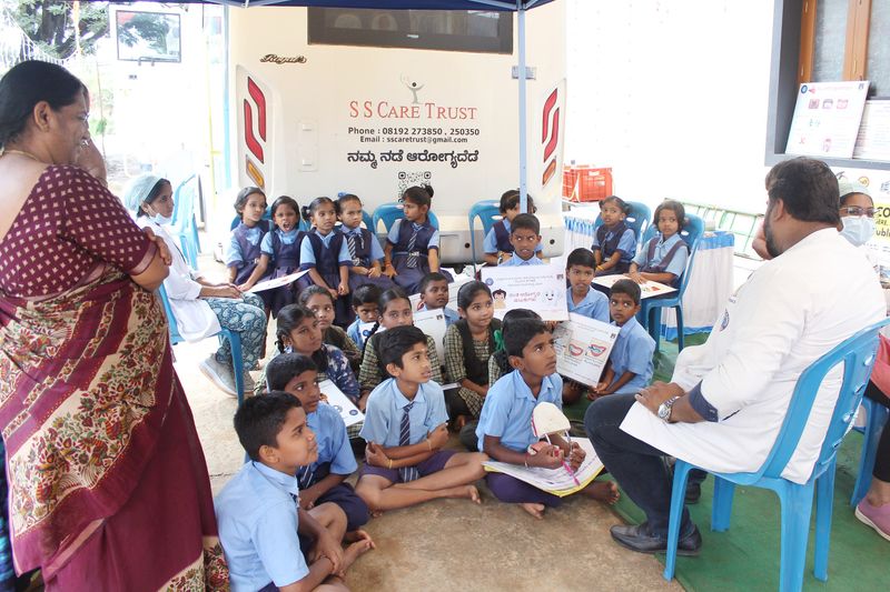 Schoolchildren sit together holding oral hygiene charts as a dentist speaks with them at an SS Care Trust health camp.