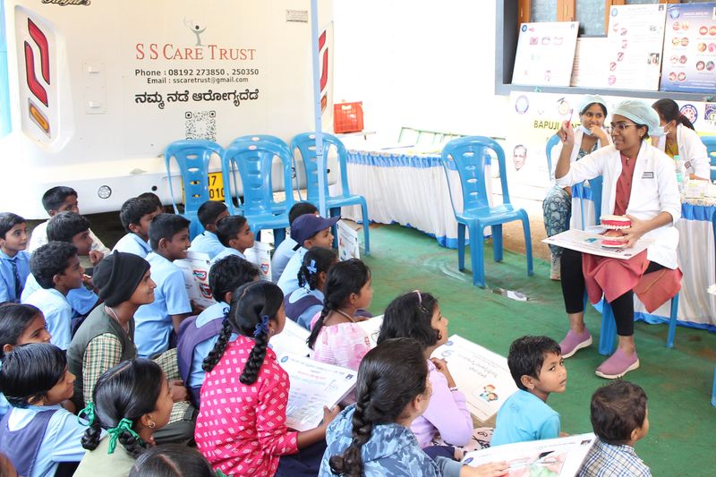 A dentist holds up a model of teeth to demonstrate brushing technique to a group of attentive schoolchildren.