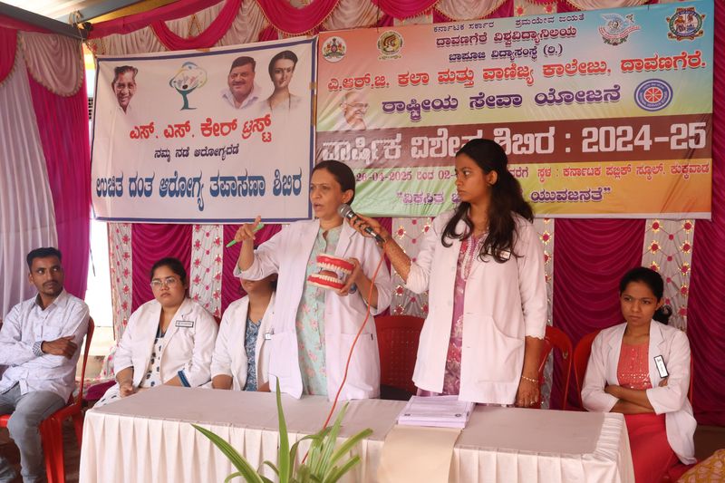 Dental college staff address an audience while holding a model of teeth at an SS Care Trust oral health camp event.