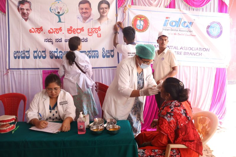 At a registration table, a staff member fills out forms as a masked dentist examines a seated woman nearby.