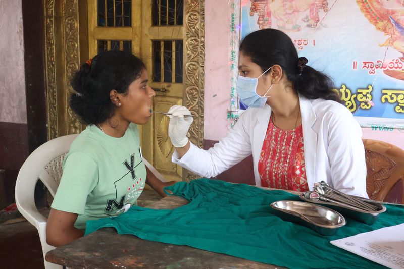 A masked health worker examines a schoolgirl's mouth with a dental instrument at an oral health screening camp.