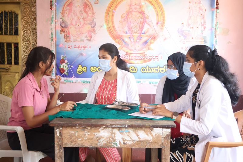 Four masked dental camp volunteers in white coats confer around a table during a school oral health camp.