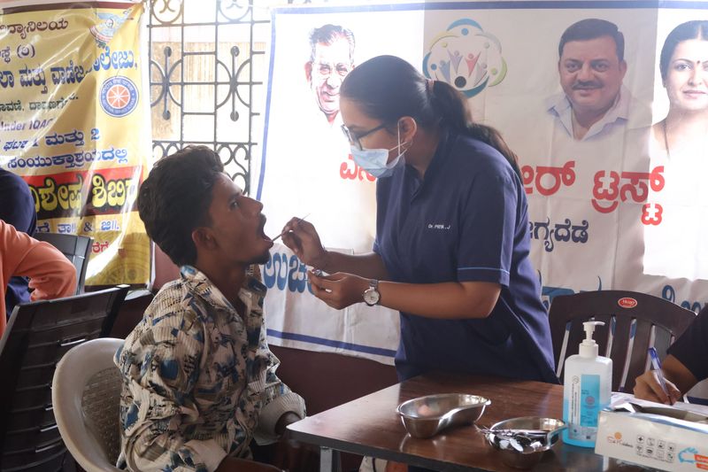 A masked dental worker in scrubs examines a young patient's mouth at an outdoor oral health screening camp.