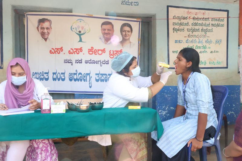 A masked dental worker examines a young woman's mouth beside an SS Care Trust free dental camp banner.