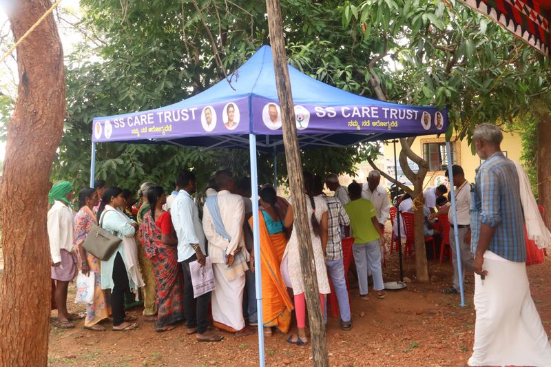 A crowd of people gathers under an SS Care Trust tent waiting for health camp services beneath shady trees.