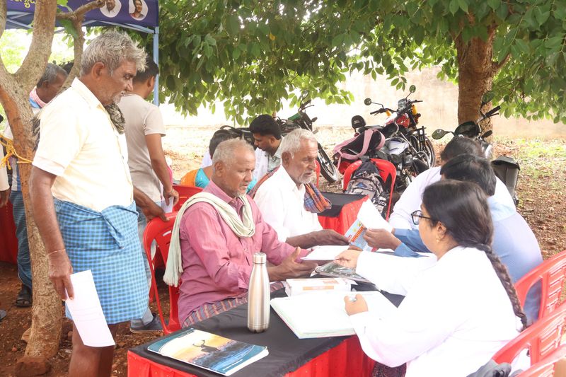 Volunteers hand educational booklets to seated men at an outdoor community health camp table.
