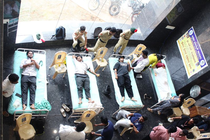 An overhead view of a blood donation camp with donors resting on cots and uniformed volunteers nearby.