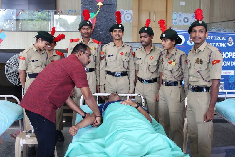 NCC cadets in ceremonial uniform gather around a blood donor on a cot while a volunteer checks his pulse.