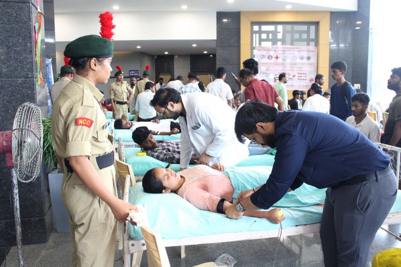 An NCC cadet looks on as a doctor and volunteer attend to a blood donor resting on a cot.