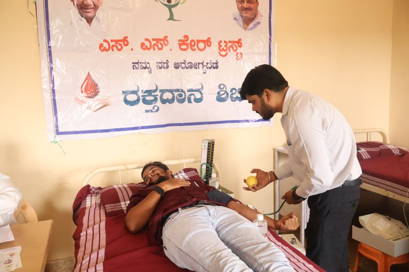 A volunteer offers a glucose drink to a blood donor resting after donating, beside an SS Care Trust camp banner.