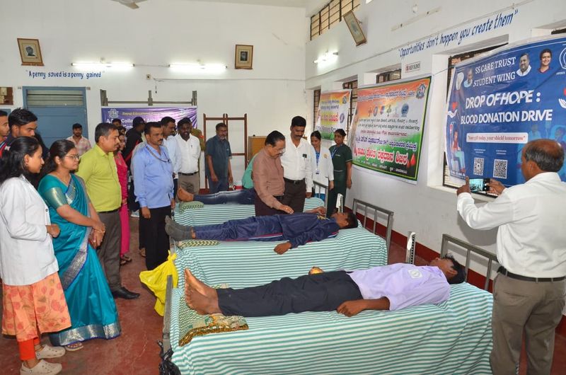 Guests and organisers walk past a row of blood donors resting on cots during a campus donation drive.