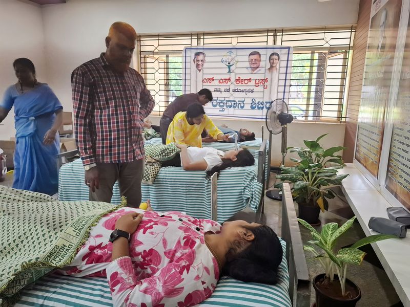 A blood donor rests on a cot in the foreground while volunteers attend to other donors at an indoor camp.