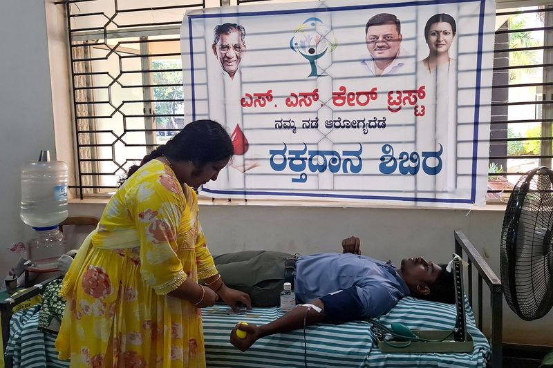 A health worker attends to a young man donating blood on a camp bed below the SS Care Trust blood donation banner.
