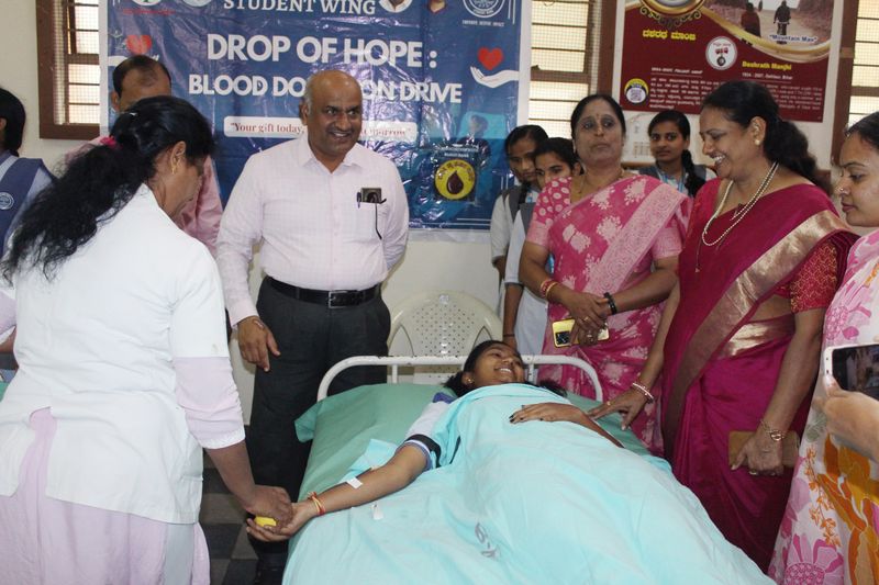 A young woman smiles while donating blood on a camp bed, surrounded by well-wishers under a Drop of Hope banner.