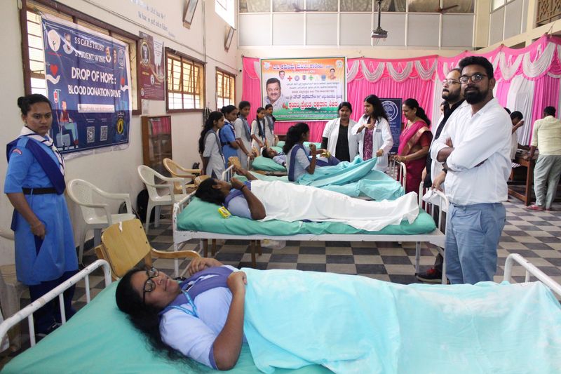 Several young women donate blood on camp beds as staff and classmates look on beneath a Drop of Hope banner.