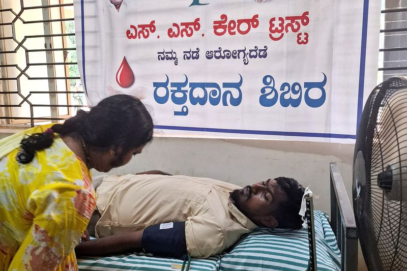 A health worker checks on a blood donor lying on a camp bed beneath the SS Care Trust blood donation banner.