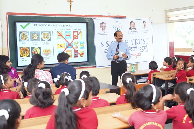 A speaker stands beside a screen titled "Let's choose healthy snacks", comparing home-made snacks with packaged foods, in front of young students in red uniforms.