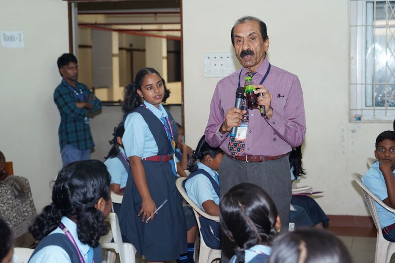 A speaker holding a microphone and a drink bottle talks to students in blue uniforms, one of whom stands to answer.