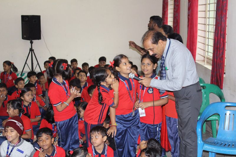 A speaker holds a microphone out to a smiling schoolgirl in a red uniform while classmates gather around in a school hall.