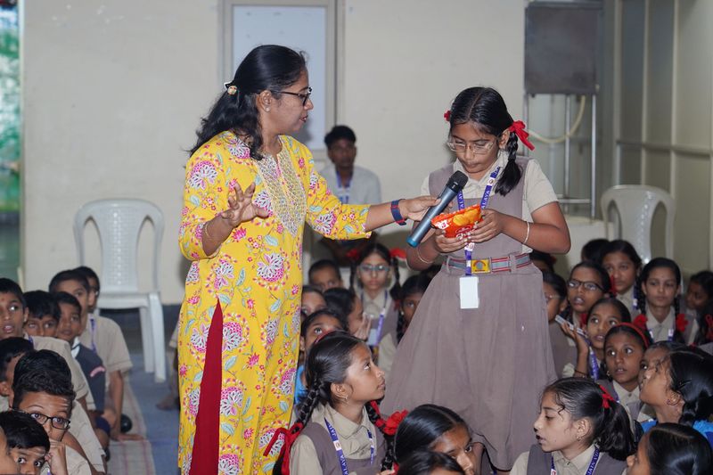 A woman facilitator talks with a schoolgirl who holds a microphone and a packet of chips during a lesson on reading snack labels.
