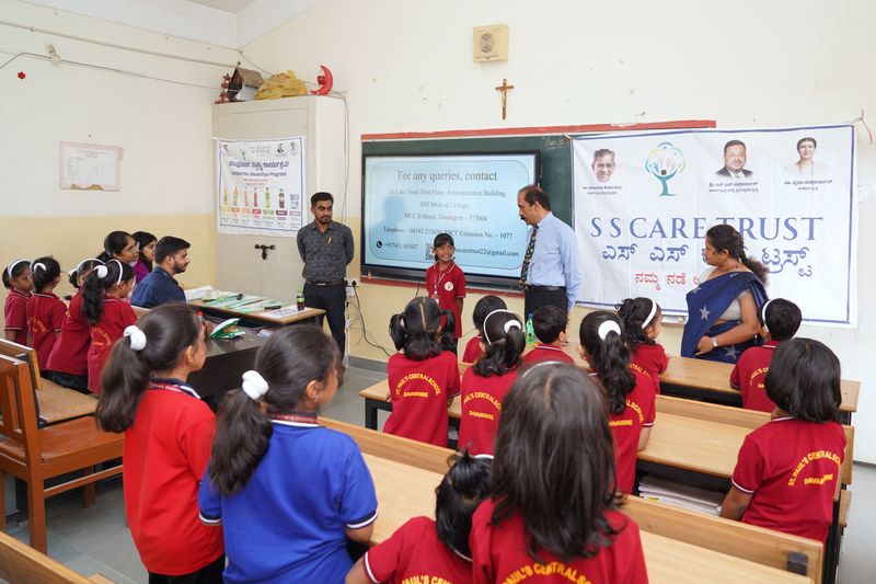 Students in red uniforms stand at their desks in a classroom with an SS Care Trust banner and the Trust's contact details on screen.