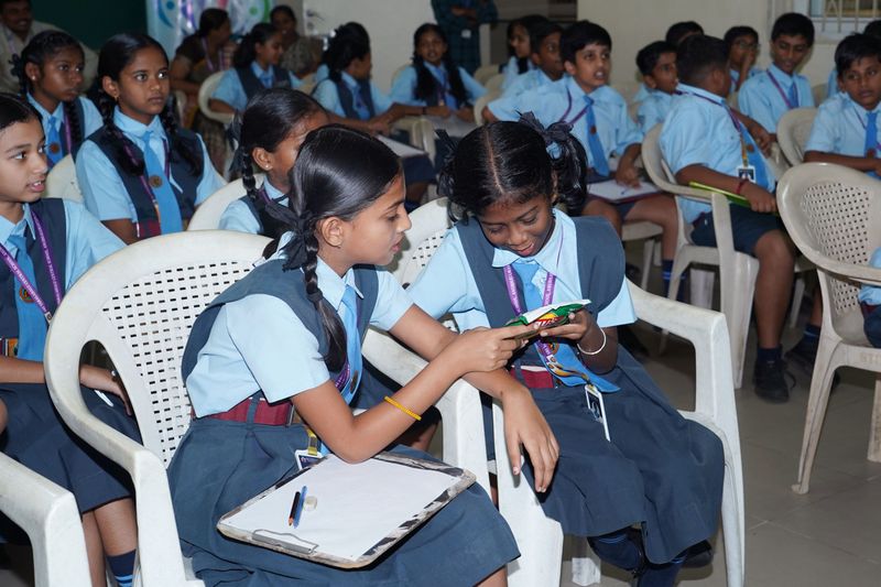Two schoolgirls in blue uniforms look closely at a snack packet together during a label-reading activity.