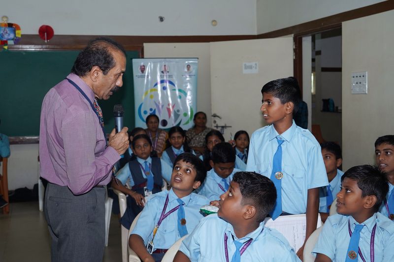 A speaker with a microphone talks with a standing schoolboy while classmates in blue uniforms listen, with an SS Care Trust standee behind.