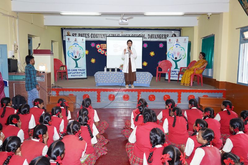 A woman in a white coat speaks from the stage to schoolgirls seated on the floor of a school hall, flanked by SS Care Trust standees.