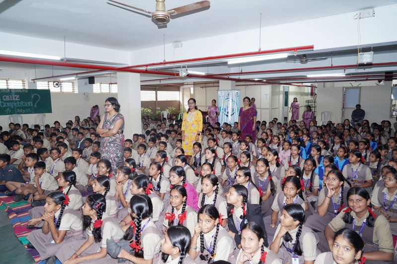 A large group of students in uniform sits on the floor of a school hall with teachers standing among them.