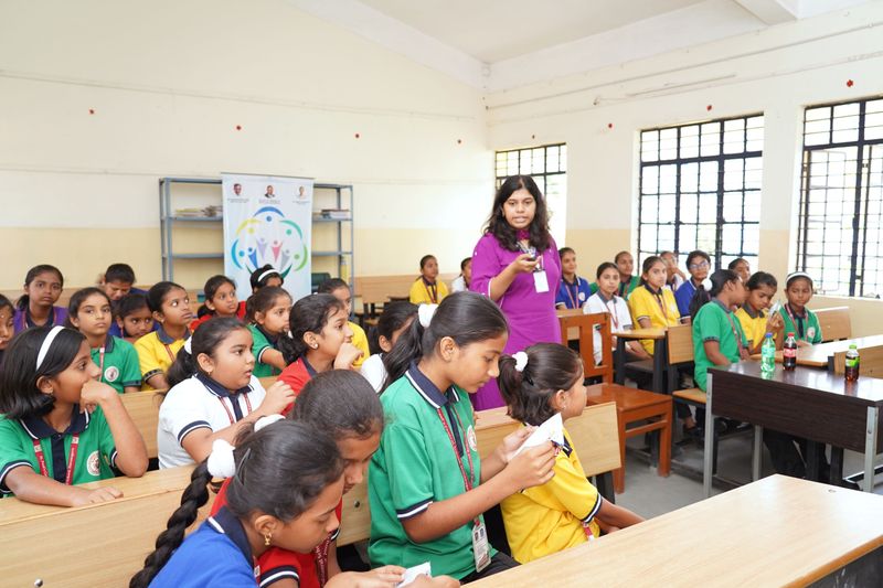 A woman facilitator walks among schoolchildren in a classroom as they work with paper handouts.