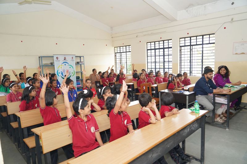 Young students in red uniforms raise their hands eagerly in a classroom while facilitators sit at a desk nearby.
