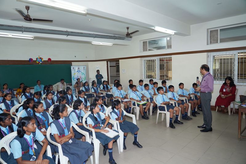 A speaker addresses rows of students in blue uniforms seated in a school hall.