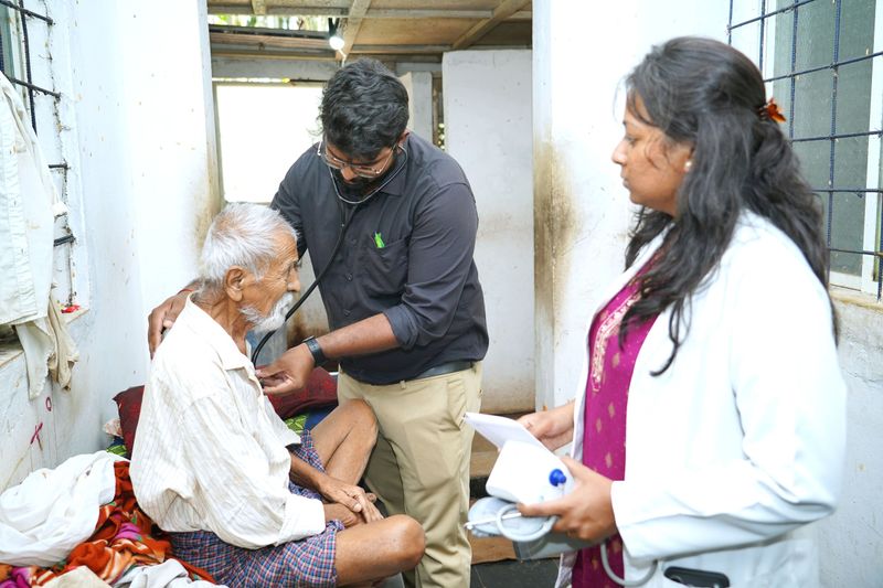 A health worker uses a stethoscope to examine an elderly man seated at home, while a colleague holds a blood pressure monitor nearby.