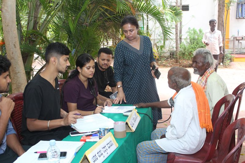 Staff check an elderly man's blood pressure and note details at an outdoor table during a community health camp, with others seated nearby.