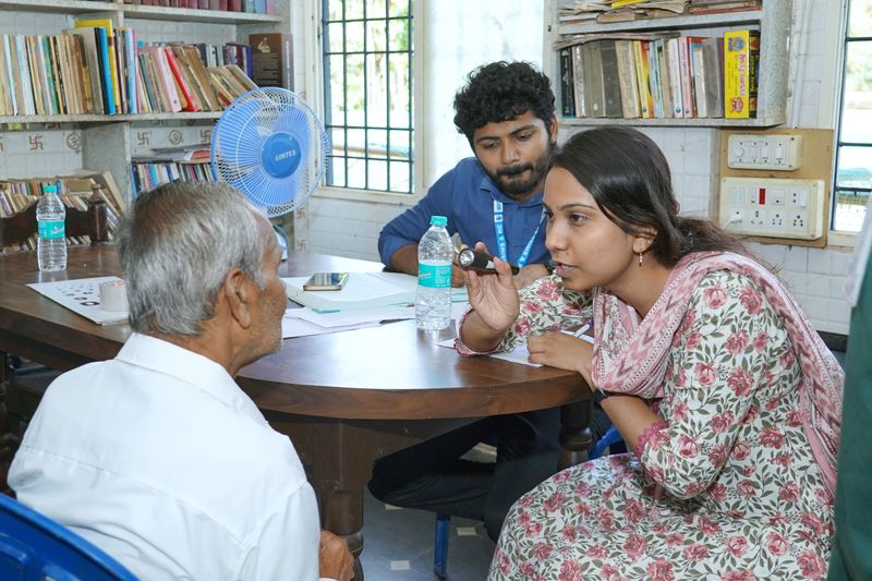An elderly man sits at a table talking with two staff members in a room lined with bookshelves, during a care visit.
