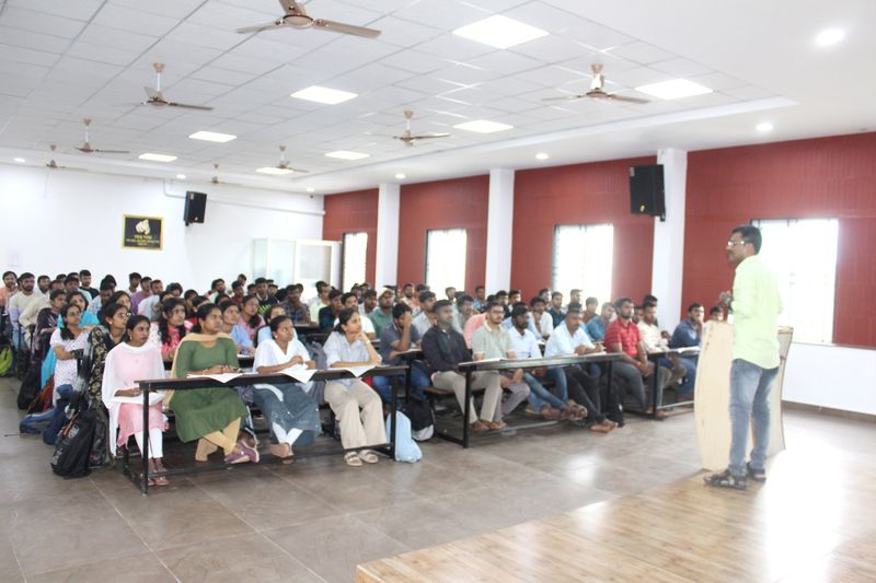 An instructor addresses a hall full of young adult students seated at long desks during a Sankalpa coaching session.