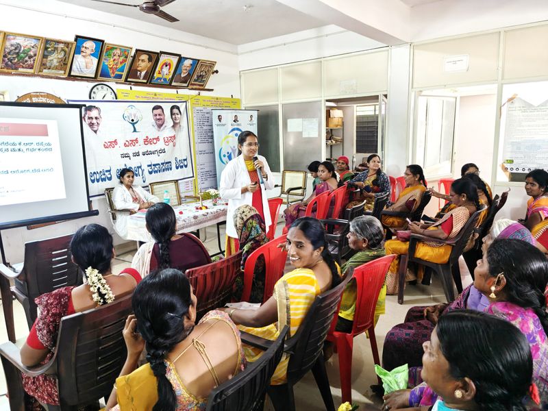 A doctor speaks to a seated group of women during a community health awareness session indoors.