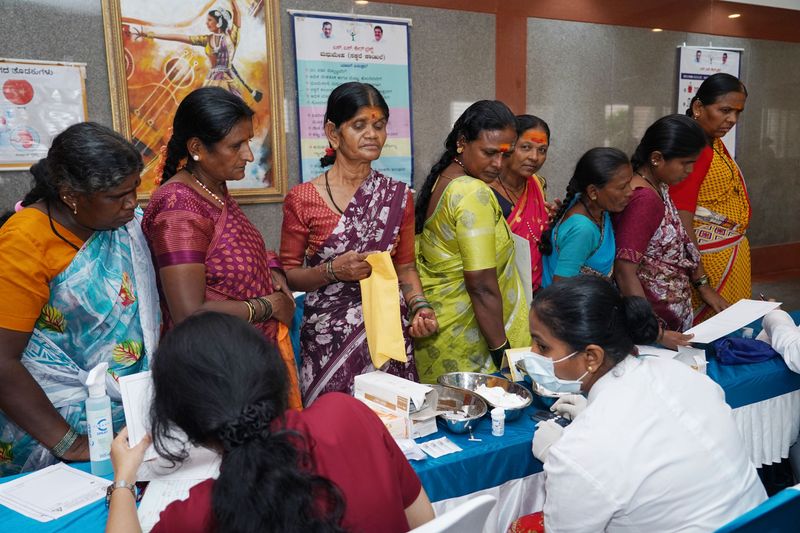 Health workers run a women's screening camp, with women queued at a table as samples are prepared.