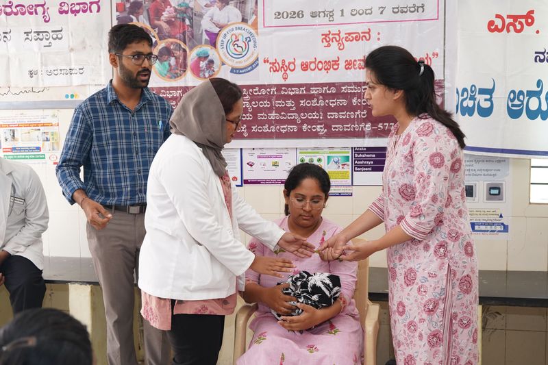 Health workers and a family member attend to a seated young woman holding a wrapped bundle, near breastfeeding-awareness banners.