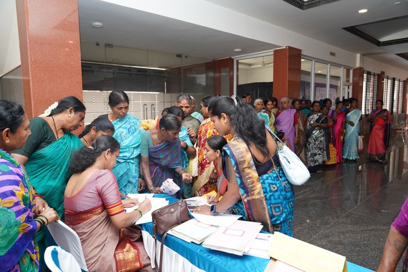 Women of different ages queue indoors with documents at a health camp registration table.