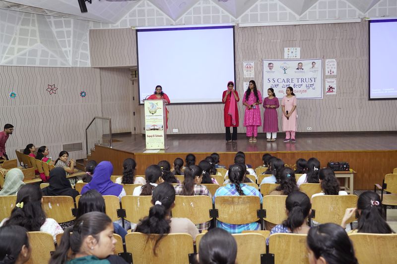 A speaker and four young women stand on stage addressing student attendees at a school health talk.