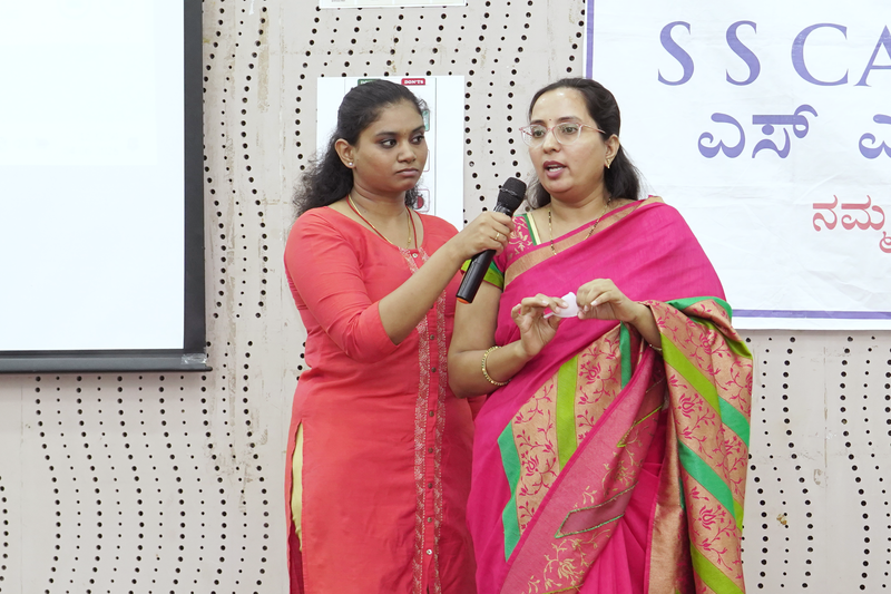 A health worker holds a microphone for a woman speaking during a community women's health awareness talk.