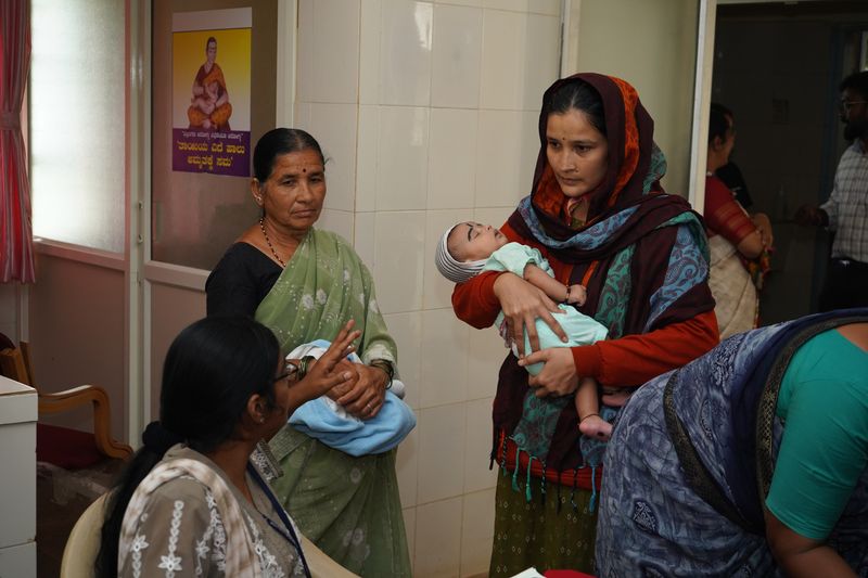A mother holds her newborn baby while a family member looks on during a postnatal care visit.