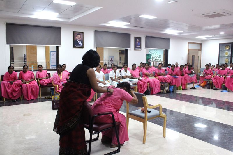 A facilitator attends to a seated woman during a health demonstration, with other women looking on.