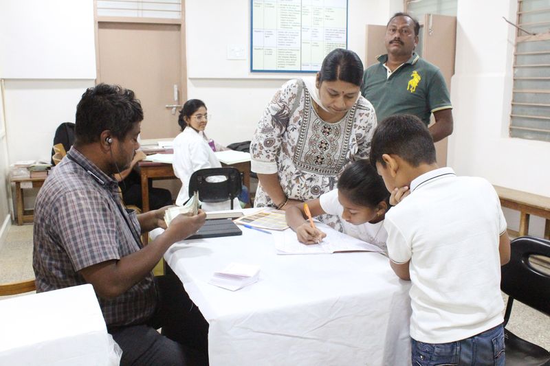 A boy and girl fill out a form at a table while staff and a seated man assist during a family health camp.