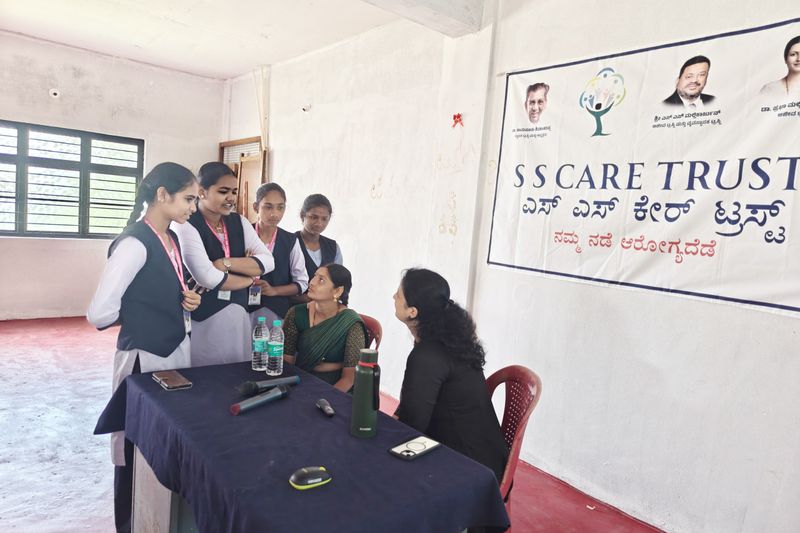 Young women in uniform talk with two seated staff members at a table in front of an S S Care Trust banner.