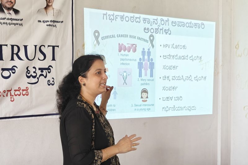 A presenter gestures while explaining cervical cancer risk factors shown on a projector screen in Kannada.