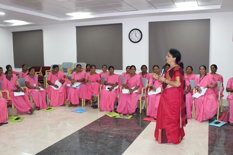 A presenter addresses rows of seated women in pink sarees during a women's group health education session.