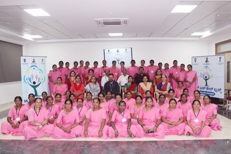 A large group of women in pink sarees pose together for a formal photo beside S S Care Trust banners.