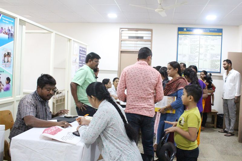 Staff register a family at a desk during a health camp, with a vaccination notice board behind them.